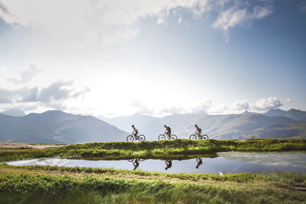 Three mountain bikers ride past a mountain lake with a panoramic view