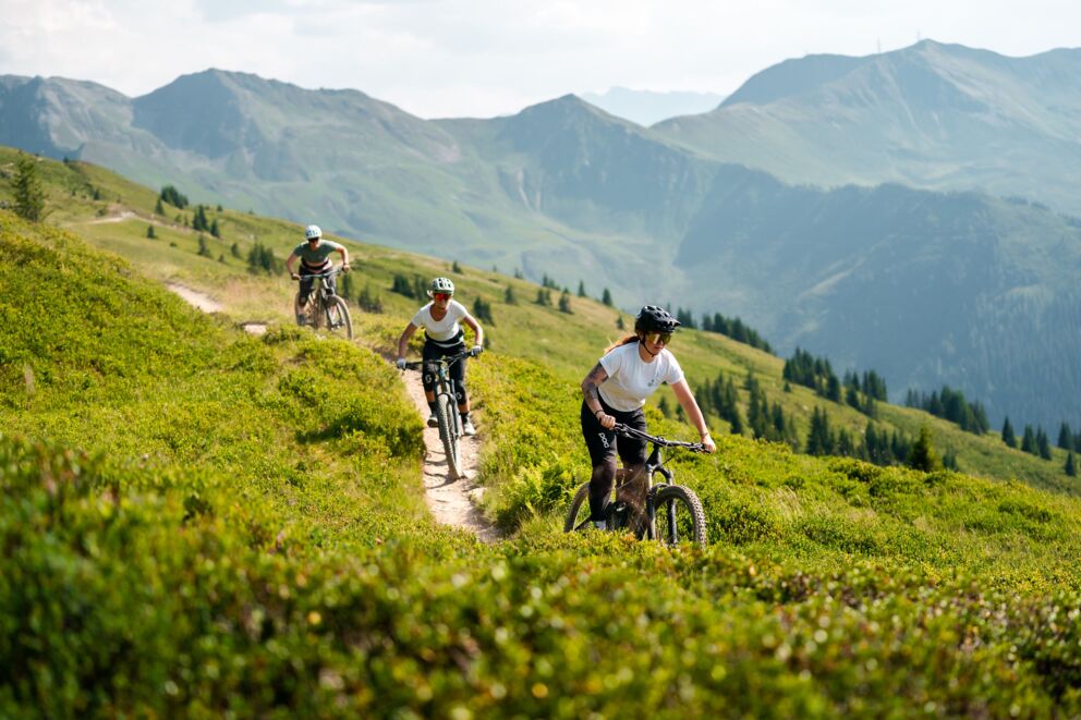 Three mountain bikers ride a trail in a green mountain scenery