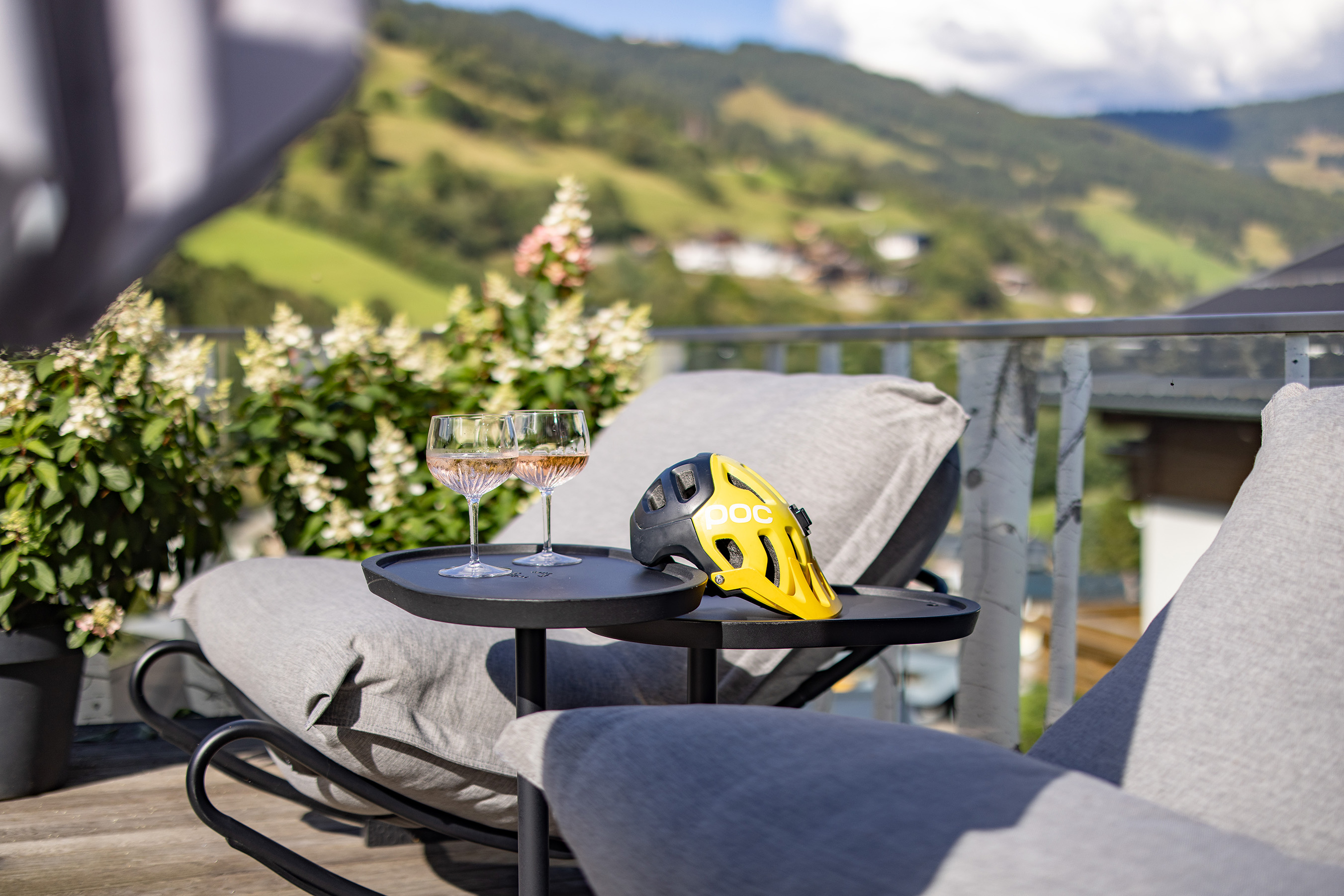 Two wine glasses and a yellow bicycle helmet on a table with mountains in the background