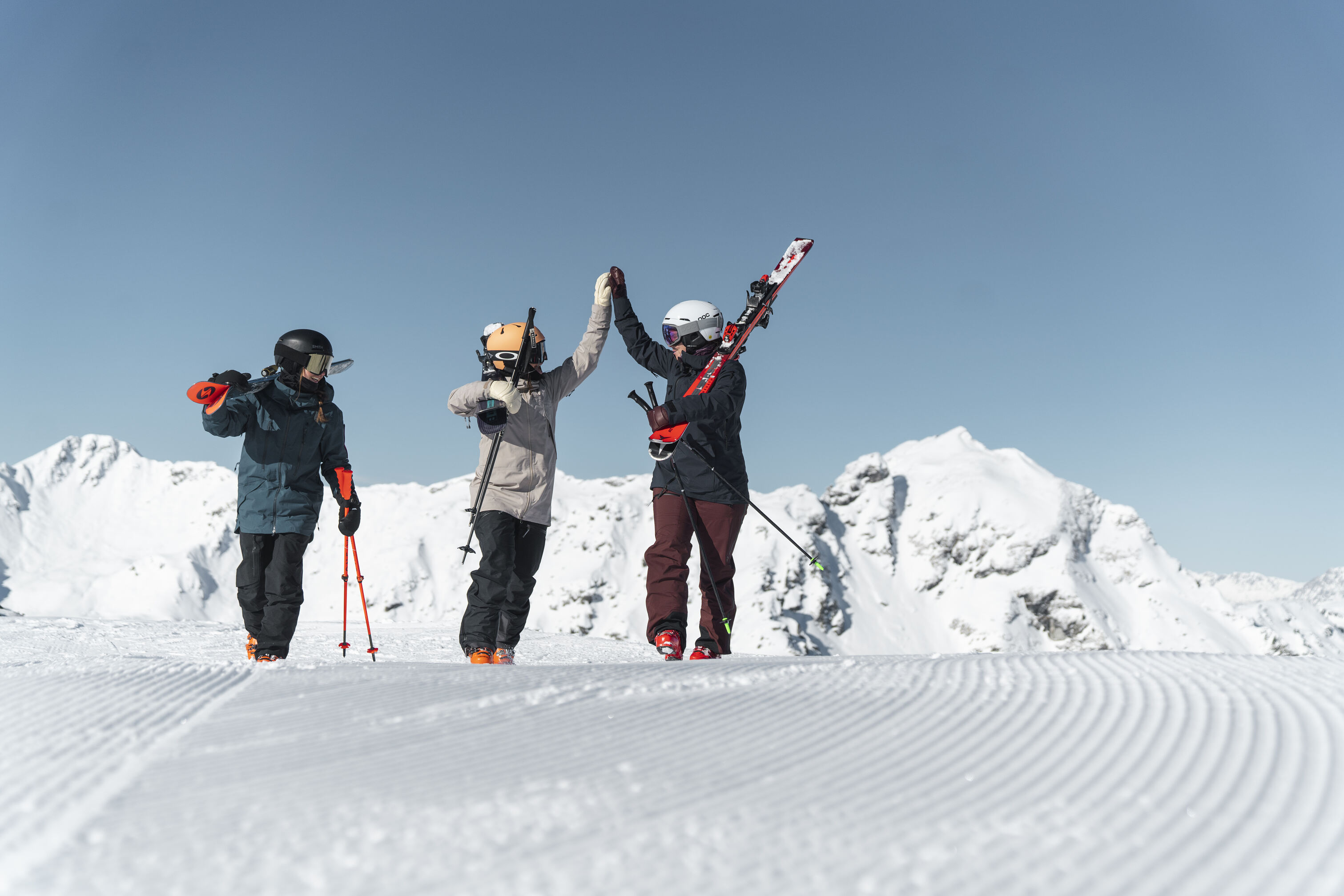 Drei Skifahrer mit geschulterten Ski vor verschneiten Berggipfeln klatschen ein