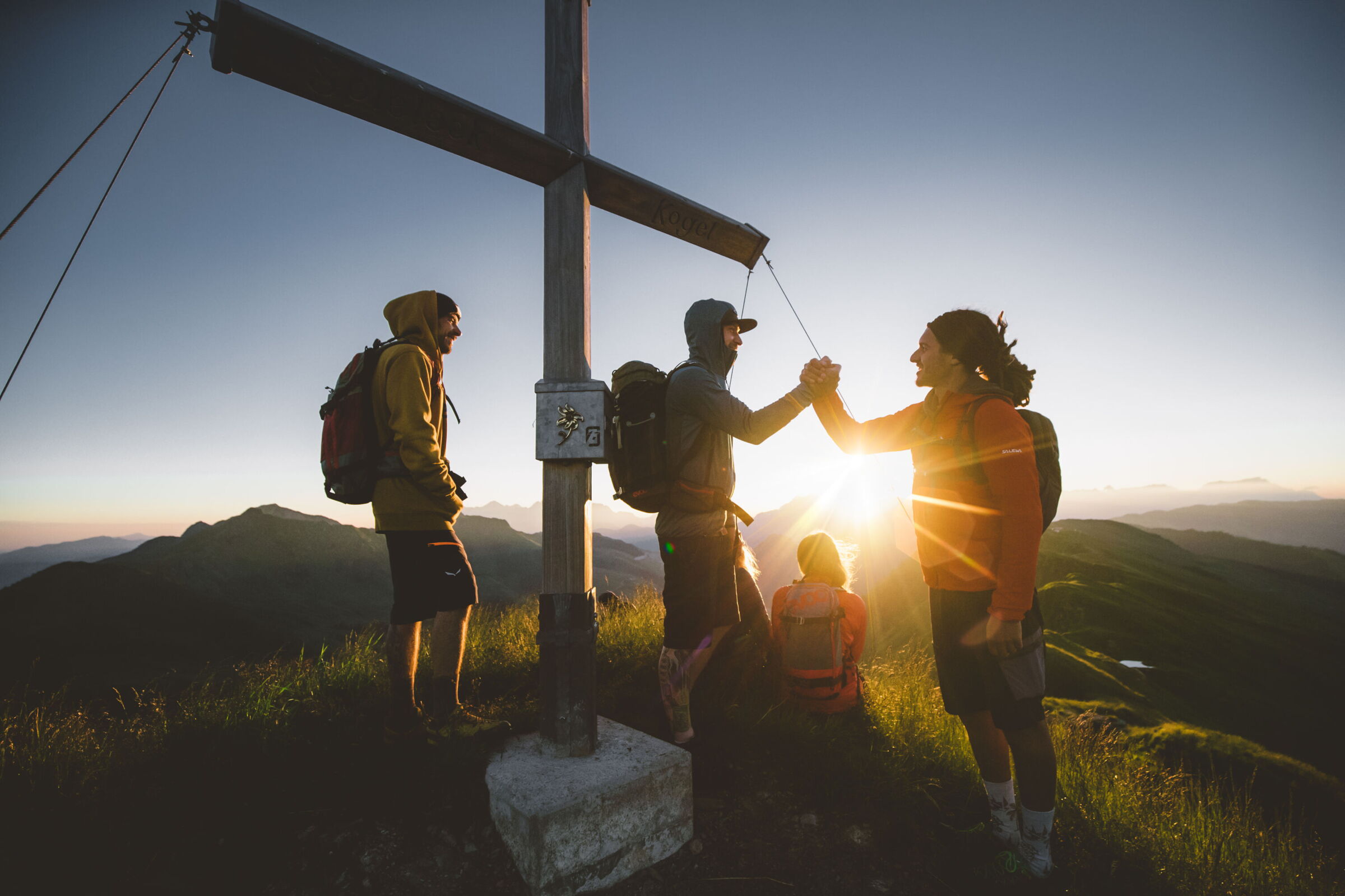 Vier Freunde stehen bei Sonnenaufgang an einem Gipfelkreuz bei einem Wanderurlaub in Hinterglemm