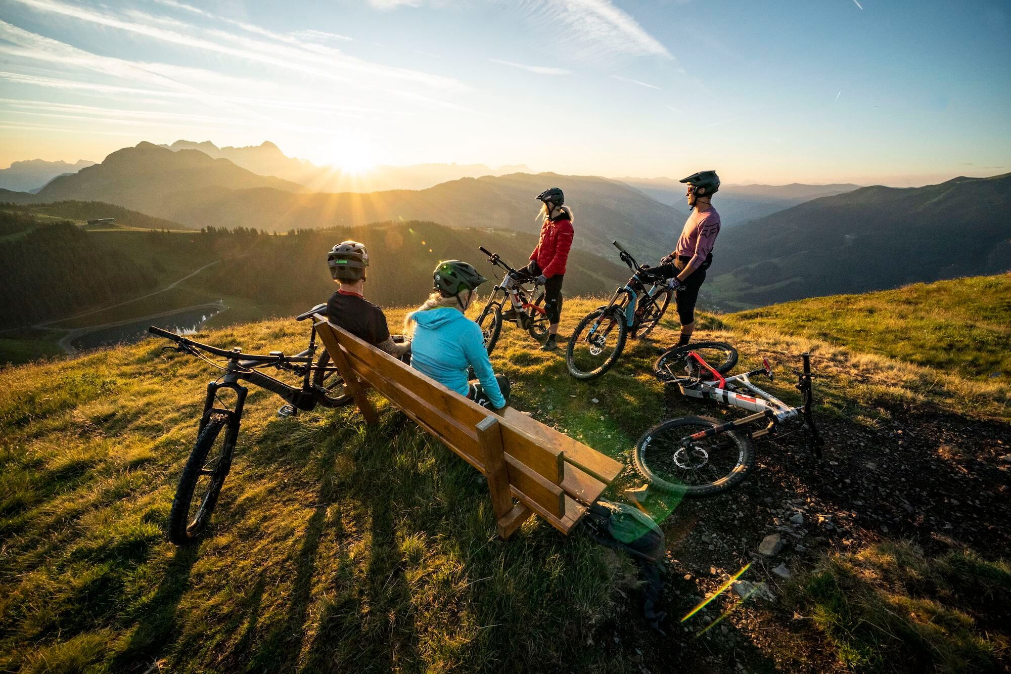 Zwei Mountainbiker machen auf einer Bank eine Pause, zwei weiterer sitzen noch auf dem Mountainbike und alle vier bestaunen den Ausblick in Saalbach